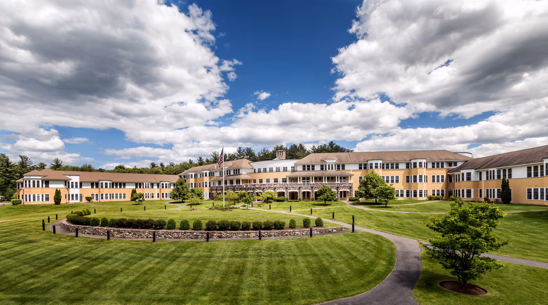 Wide exterior view of Nichols Village senior living facility with a large, well-maintained lawn, circular garden with shrubs, paved walkways, and an American flag in front of a multi-wing building under a partly cloudy sky.