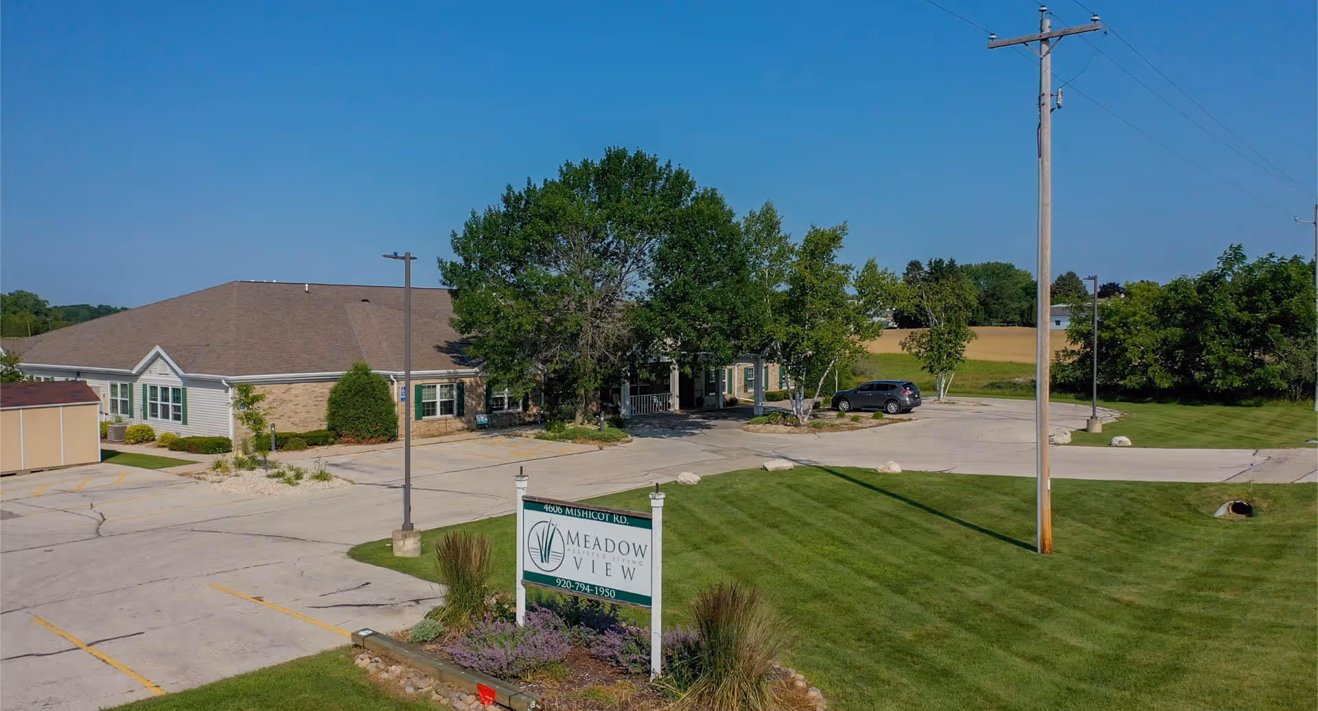 Exterior view of Meadow View Assisted Living facility showing a single-story building with a pitched roof, surrounded by trees and greenery. There is a parking lot with a few cars and a sign in the foreground displaying the facility's name and contact number. The sky is clear and blue.