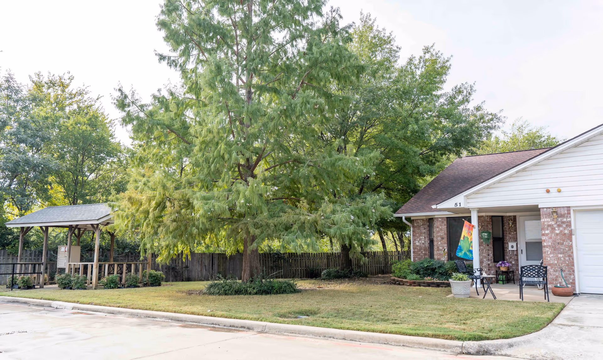 Exterior view of a single-story brick and white siding building with a small covered porch area, two black metal chairs, a small table, and a colorful flag hanging near the entrance. The building is surrounded by a grassy lawn, large green trees, and a wooden fence in the background. There is a paved driveway or parking area in the foreground.