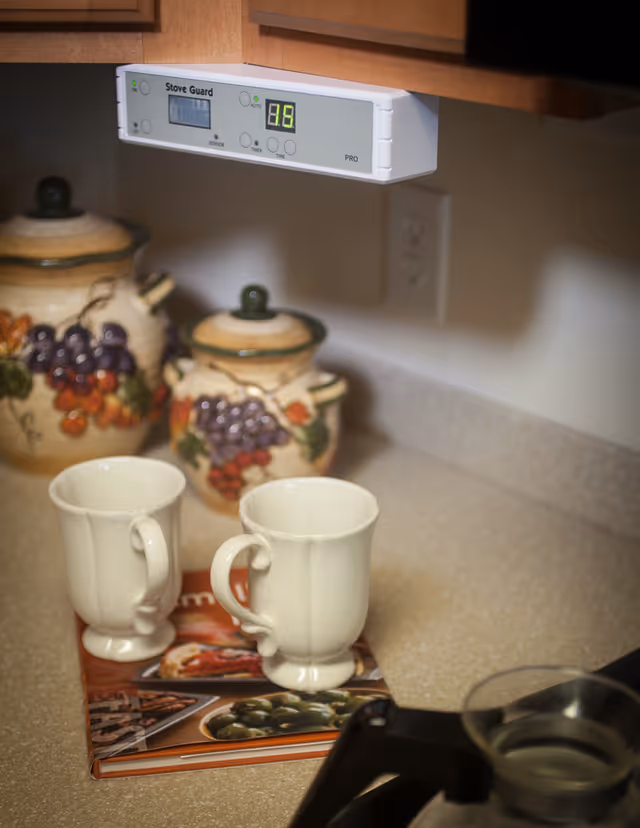 A kitchen countertop with two white ceramic mugs placed on a cookbook. Behind the mugs are two ceramic jars decorated with grapevine designs. Above the countertop is a Stove Guard device mounted under a wooden cabinet, displaying the number 16. A power outlet is visible on the wall.