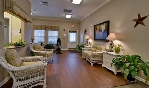 A cozy living room area in Parkway Cove featuring wicker furniture with cushions, including two armchairs and a sofa. The room has wooden flooring, potted plants, a white side table with two lamps, wall decorations including a framed picture and star-shaped ornaments, and a door with windows letting in natural light.