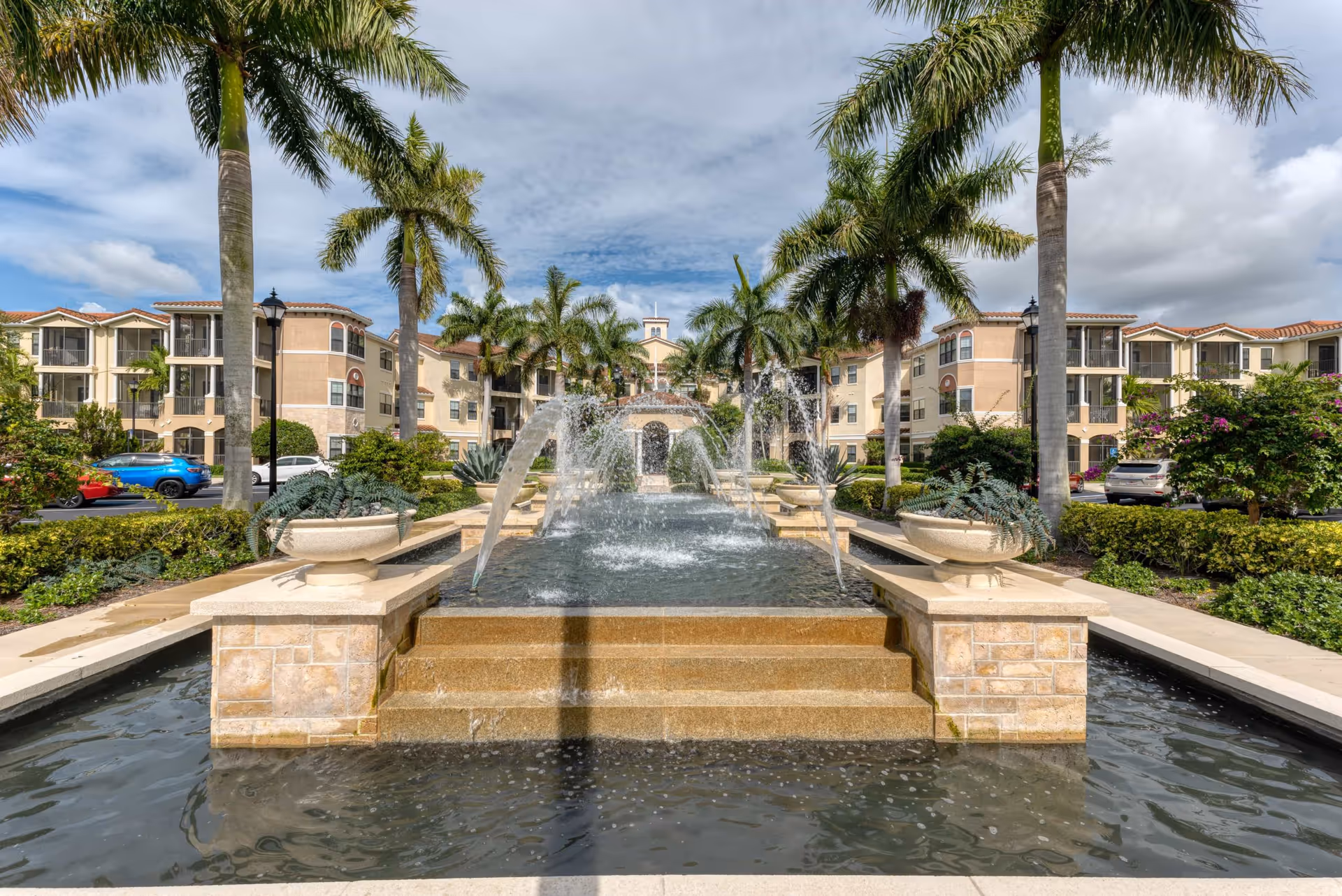 Outdoor view of Addington Place of Jupiter featuring a large decorative water fountain with multiple water jets, surrounded by palm trees and landscaped greenery, with a multi-story residential building in the background under a partly cloudy sky.
