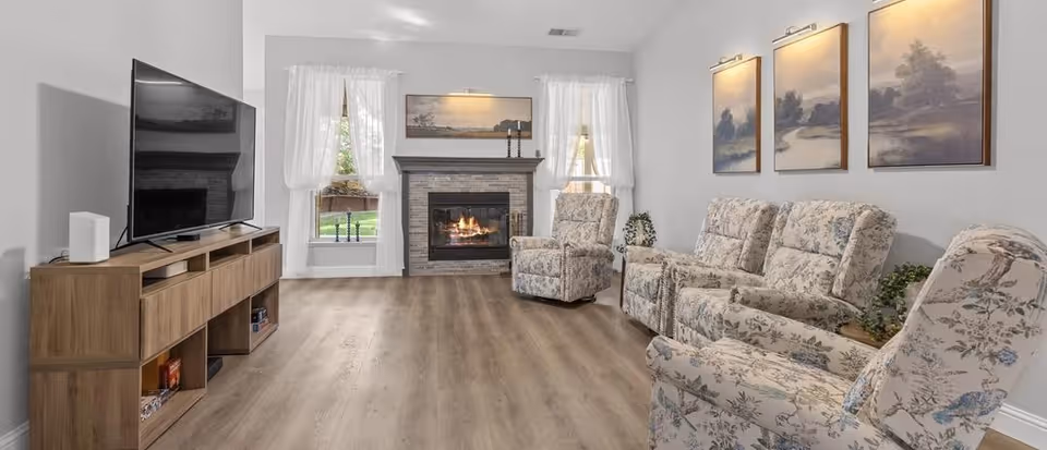 A cozy living room with a wooden floor, a fireplace with a lit fire, and a TV on a wooden stand. The room has floral patterned armchairs and a sofa, three landscape paintings on the wall, and two windows with white curtains letting in natural light.