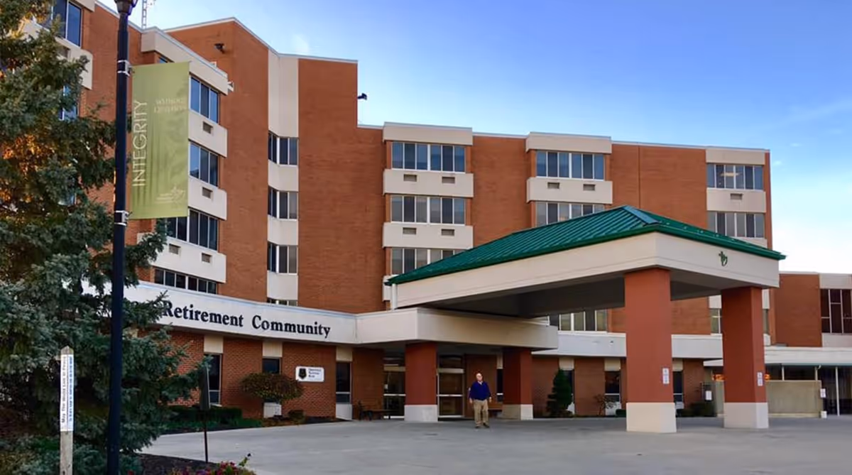 Exterior view of a multi-story brick retirement community building with a green roofed entrance canopy. A person is walking near the entrance, and there is a green banner on a lamppost that reads 'INTEGRITY'.