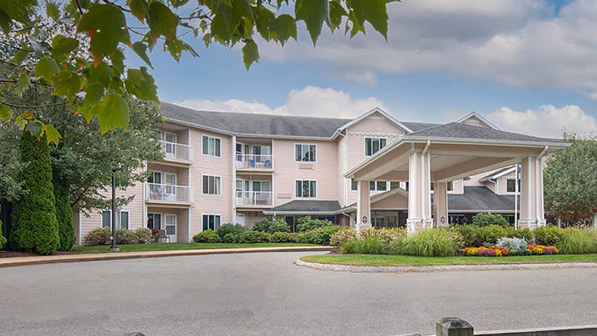 Exterior view of a three-story senior living facility building with balconies, surrounded by trees and landscaping, under a partly cloudy sky.