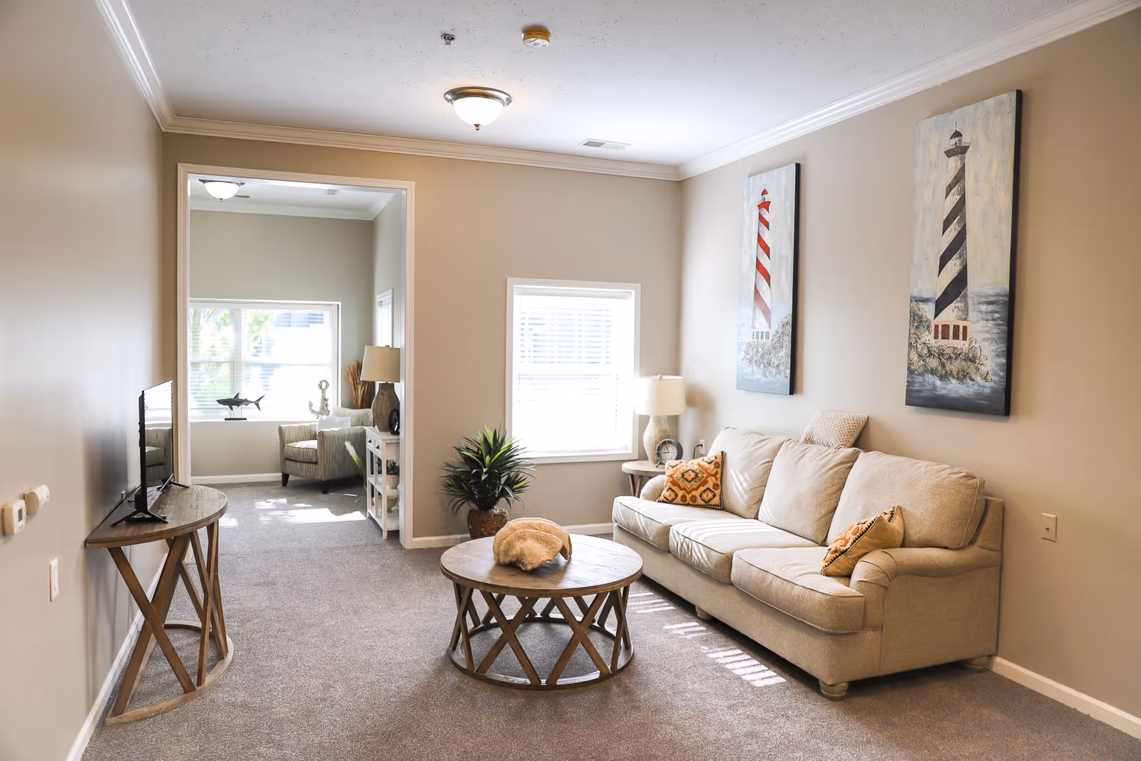 Well-lit living room with a beige sofa, round wooden coffee table, TV on a console, and lighthouse wall art.