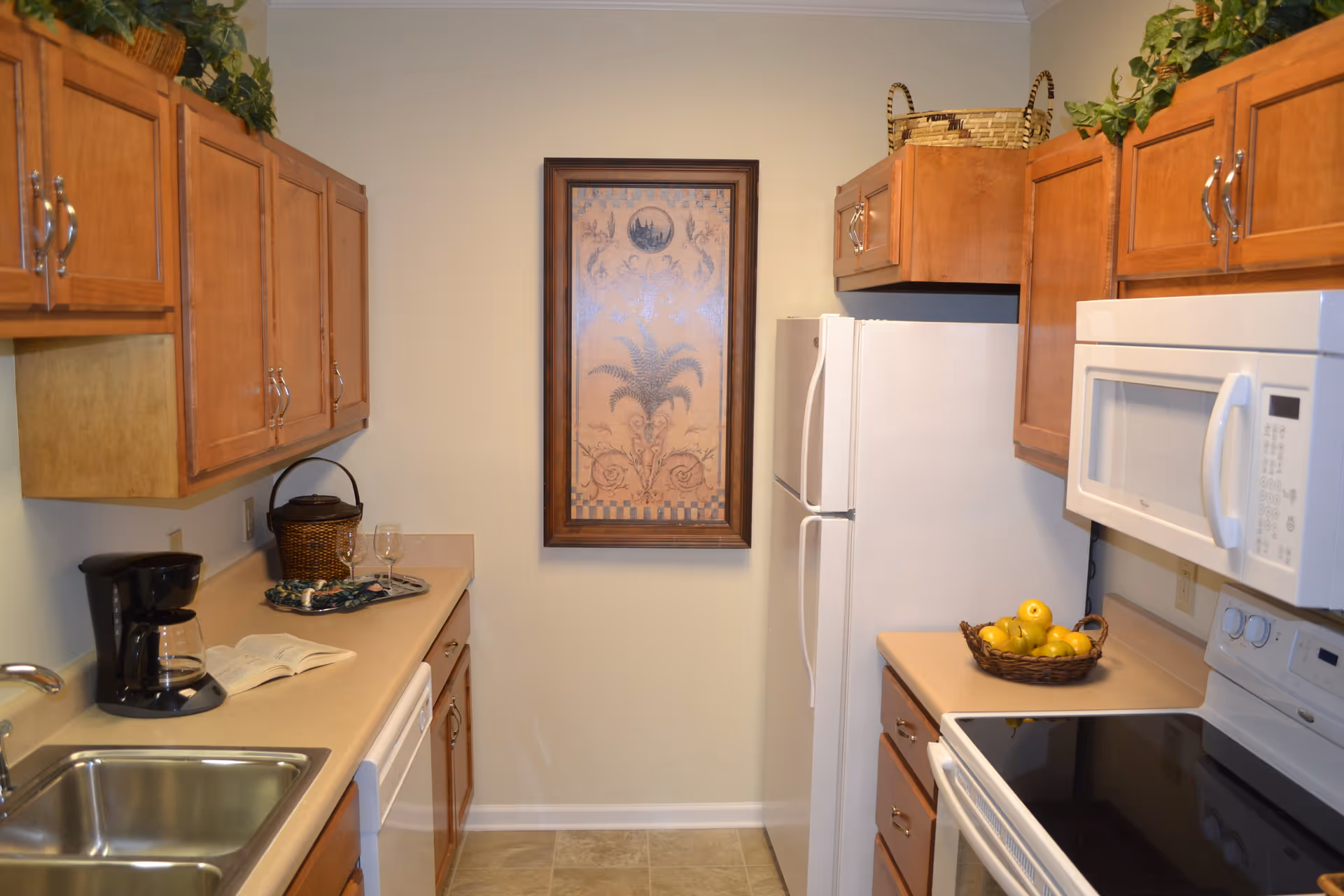 A compact kitchen with wooden cabinets, a white refrigerator, microwave, and stove. The countertop has a basket of apples, a coffee maker, a glass, and an open book. A framed decorative artwork hangs on the wall at the end of the kitchen.