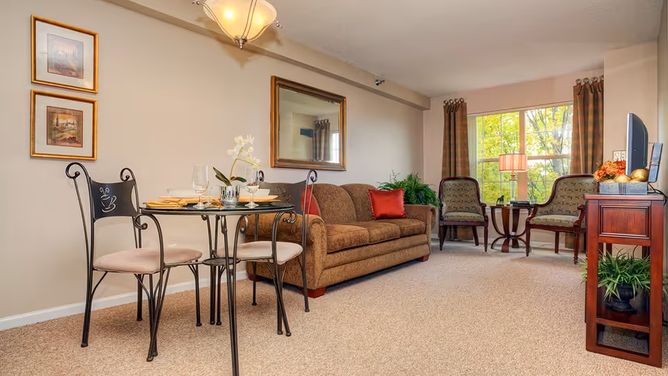 A cozy living room area with a brown sofa adorned with red pillows, two patterned armchairs near a window with brown curtains, a small round glass dining table with four chairs, and a wooden TV stand with a television and decorative items. The room is carpeted and has framed artwork on the wall and a large mirror above the sofa.