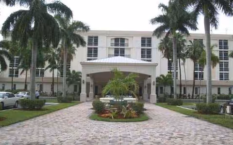 Paved driveway leads to a white, multi-story senior living facility front entrance framed by palm trees.