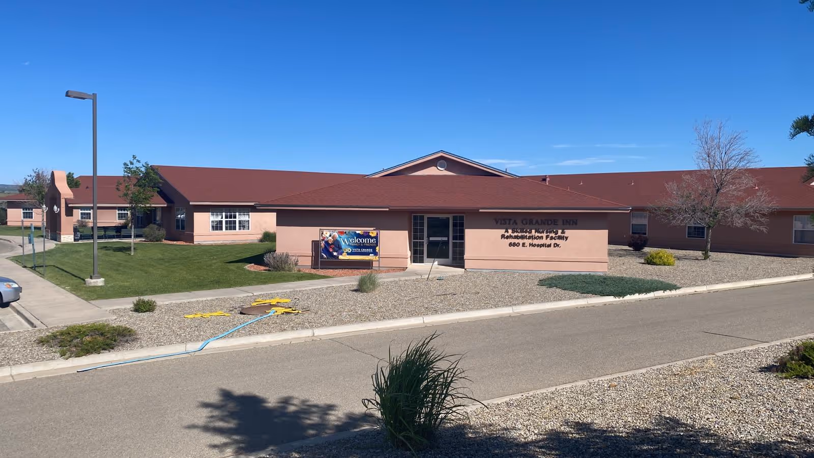 Exterior view of Vista Grande Inn Rehabilitation Facility, a single-story building with a red roof and beige walls. The building is surrounded by a landscaped area with grass, bushes, and gravel. A sign near the entrance reads 'Vista Grande Inn Rehabilitation Facility 680 E. Hospital Dr.' and another sign welcomes visitors. The sky is clear and blue.