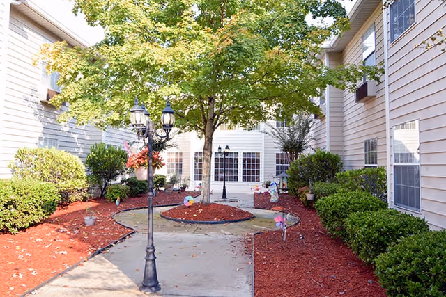 Outdoor courtyard area at Brookside Stone Mountain with a central tree surrounded by mulch, bushes along the building walls, a concrete pathway, and decorative lamp posts with hanging flower baskets.