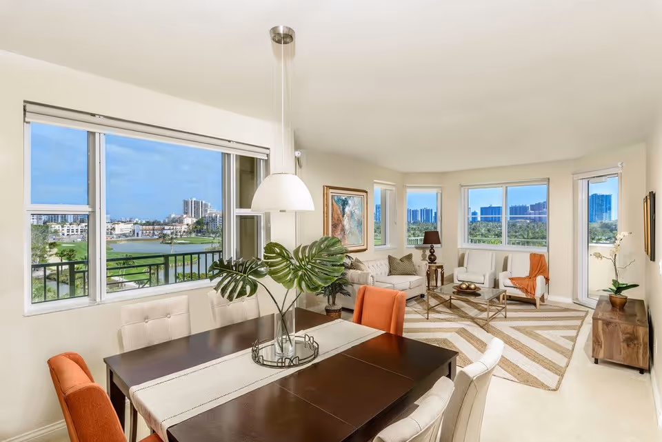 Bright and spacious living and dining area in a senior living facility with large windows offering a view of a golf course and cityscape. The dining area features a dark wooden table with a white runner, surrounded by beige and orange chairs, and a vase with large green leaves as a centerpiece. The living area includes a beige sofa, two white armchairs with an orange throw, a glass coffee table on a patterned rug, a wooden sideboard with a plant, and framed artwork on the walls.