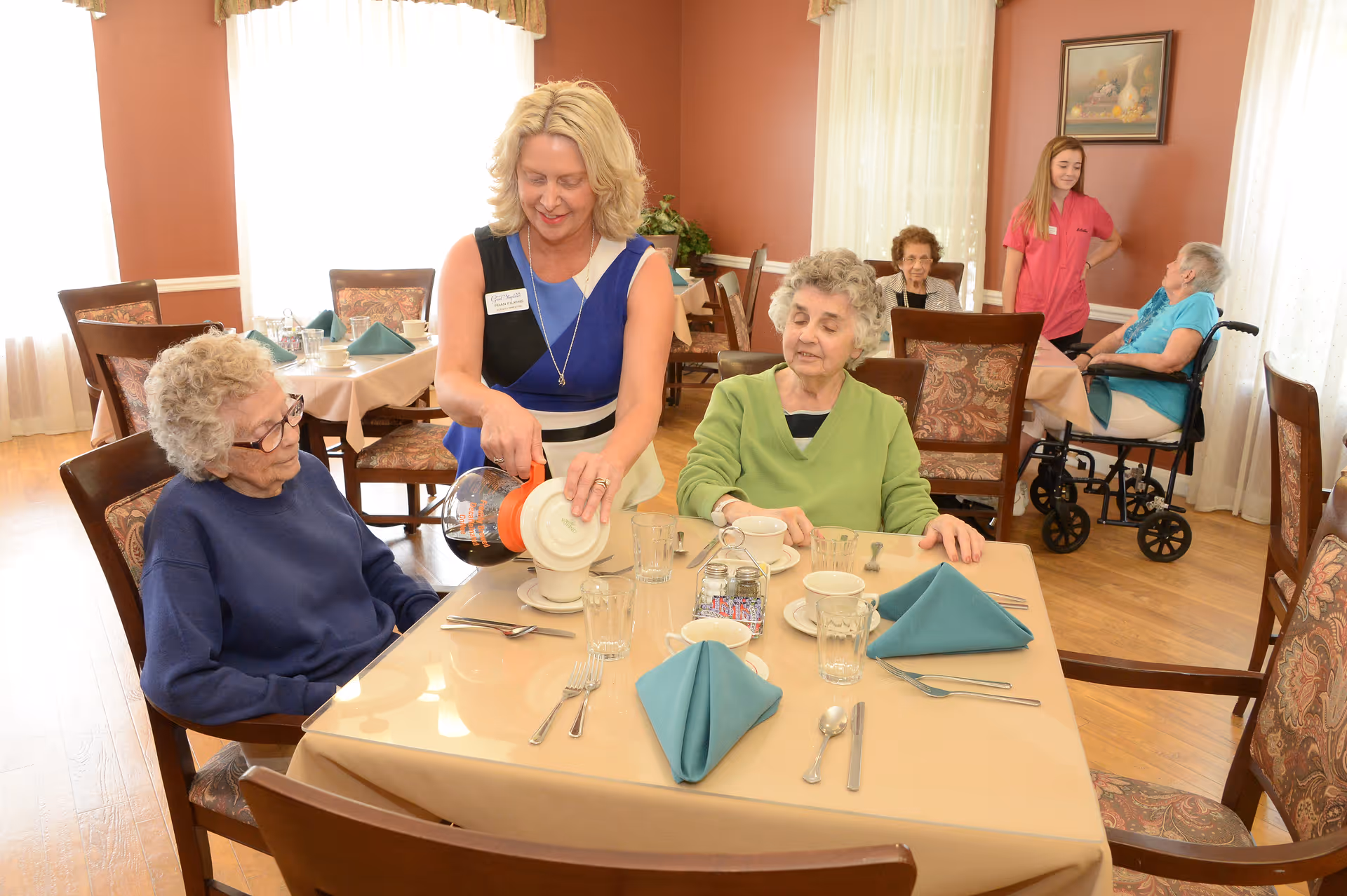 A staff member pours coffee for two elderly residents seated at a set dining table in a senior living dining room.