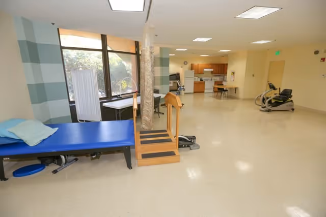 A spacious rehabilitation room with exercise equipment including a blue padded therapy table, wooden stairs for physical therapy, and a stationary exercise bike. The room has large windows letting in natural light, a kitchenette area with cabinets, a refrigerator, and a microwave in the background. The floor is light-colored and the walls are painted in soft tones.