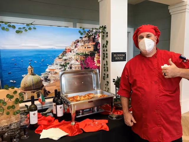 A chef wearing a red uniform, red hat, and white face mask stands next to a buffet table with a chafing dish containing food. The table is decorated with red cloth, wine bottles, glasses, and a small Italian flag. Behind the table is a large scenic backdrop of a coastal town with colorful buildings and the sea. A sign on the wall reads 'Blossom House.'
