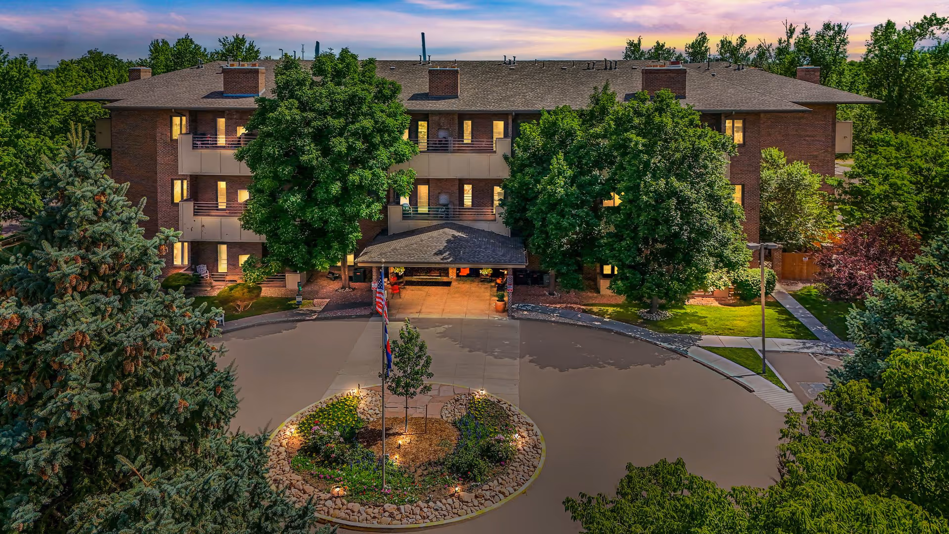 Front entrance of a three-story brick senior living building with a circular driveway, flags, and trees at dusk.