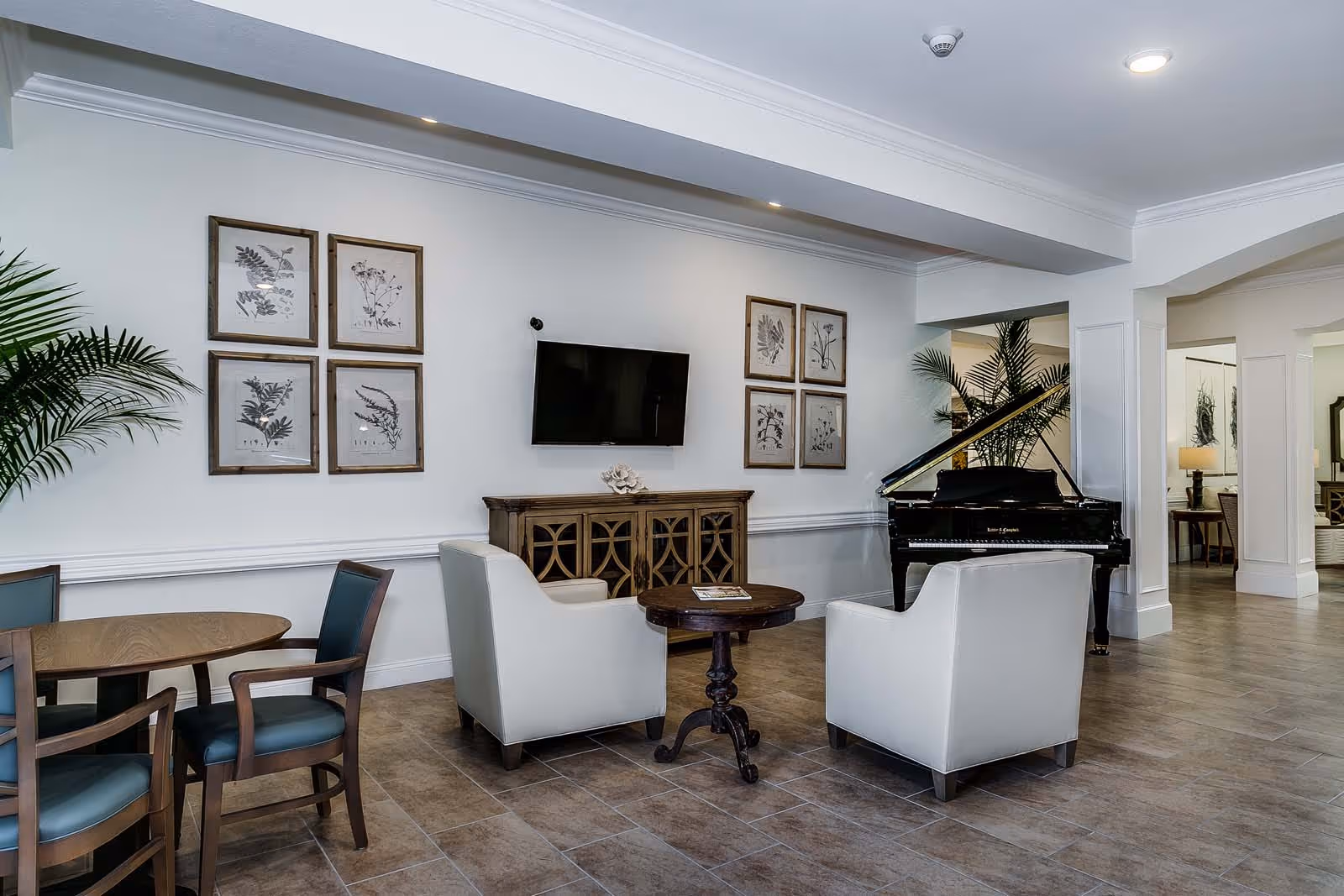 Bright common sitting area with two armchairs around a small table, a wall-mounted TV, framed botanical prints, and a black grand piano in the background.