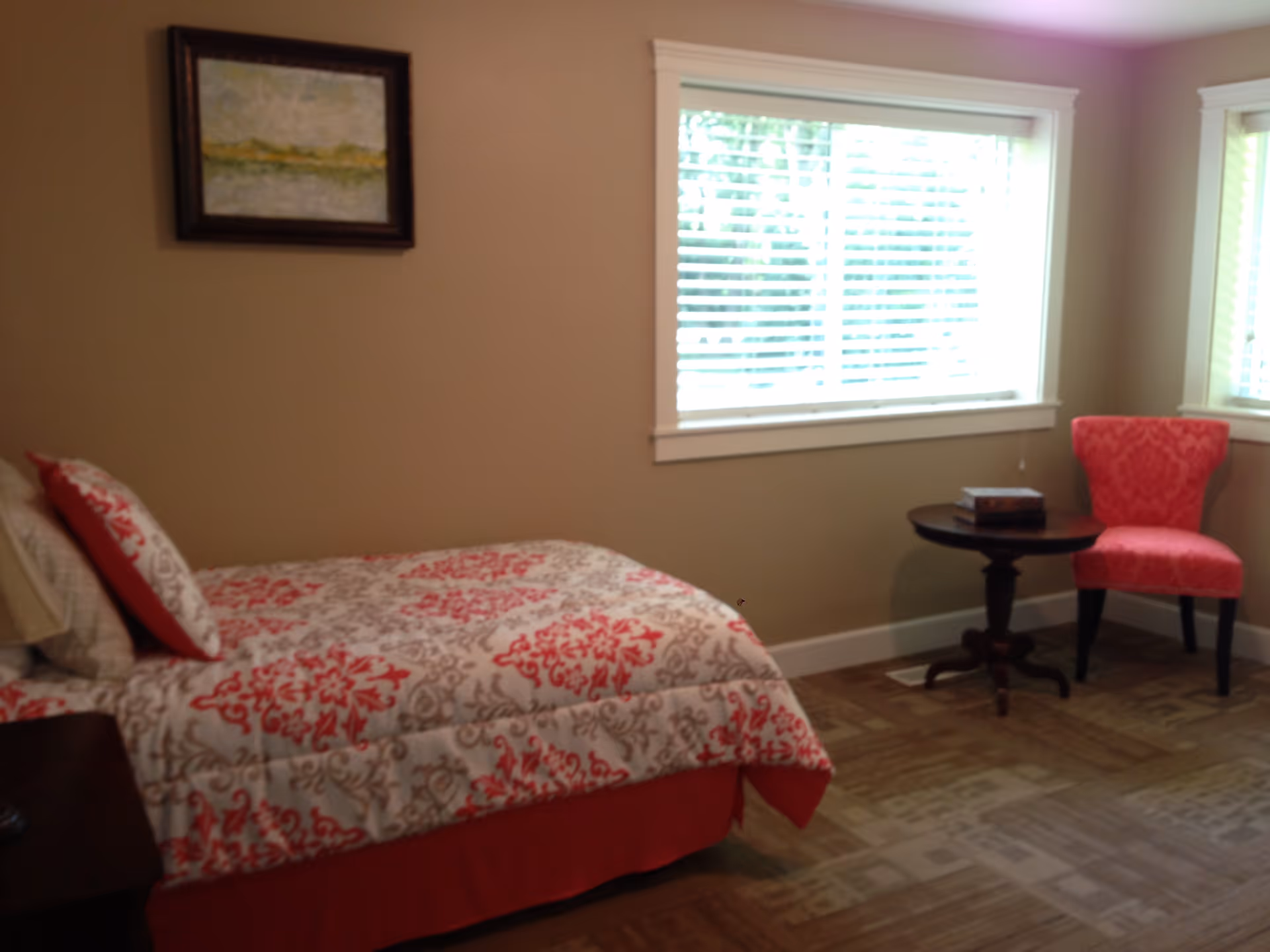 A bedroom with a single bed covered in a white and red patterned bedspread, a matching pillow, a wooden nightstand, a small round table with two books on it, a red upholstered chair, and two windows with white blinds letting in natural light. A framed painting hangs on the beige wall above the bed.