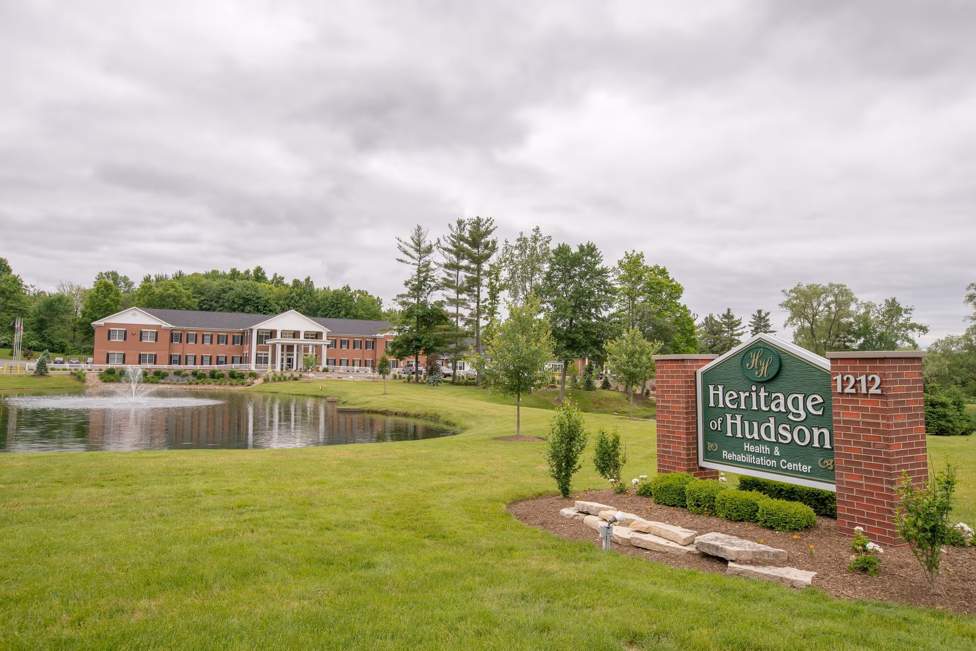 Exterior view of Heritage of Hudson Health & Rehabilitation Center showing a large brick building with white columns, a pond with a fountain in front, green lawn, trees, and a sign with the facility name and address 1212.