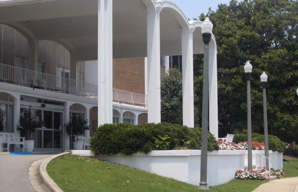 Exterior view of a building entrance with large white columns, a covered walkway, and landscaped bushes and flowers. There are several street lamps and a curved driveway leading to the entrance.