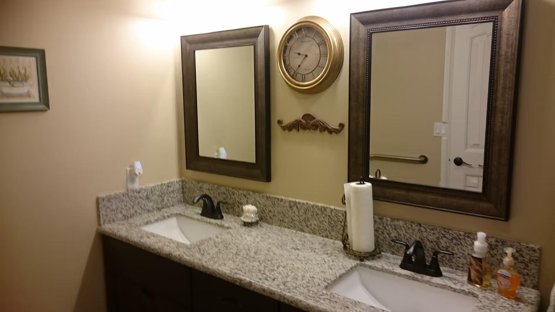 A bathroom countertop with two sinks and dark bronze faucets. Above each sink is a large framed mirror. Between the mirrors is a round wall clock with a gold frame and a decorative wooden wall accent below it. On the countertop, there is a paper towel holder, a soap dispenser, and a small decorative item. The walls are painted beige.