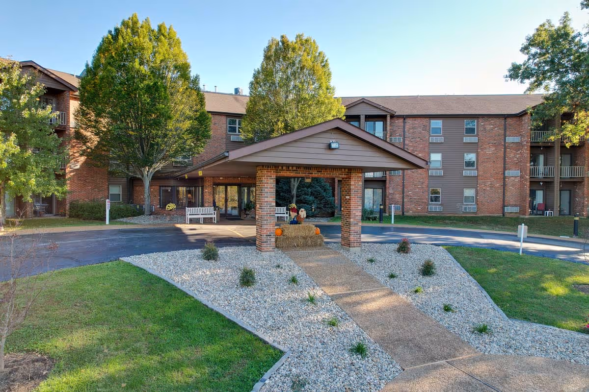 Exterior view of Autumn Ridge Residences, showing a brick building with multiple windows and balconies. In front, there is a covered entrance supported by brick pillars, surrounded by landscaped areas with grass, small plants, and white gravel. Trees are visible around the building under a clear blue sky.