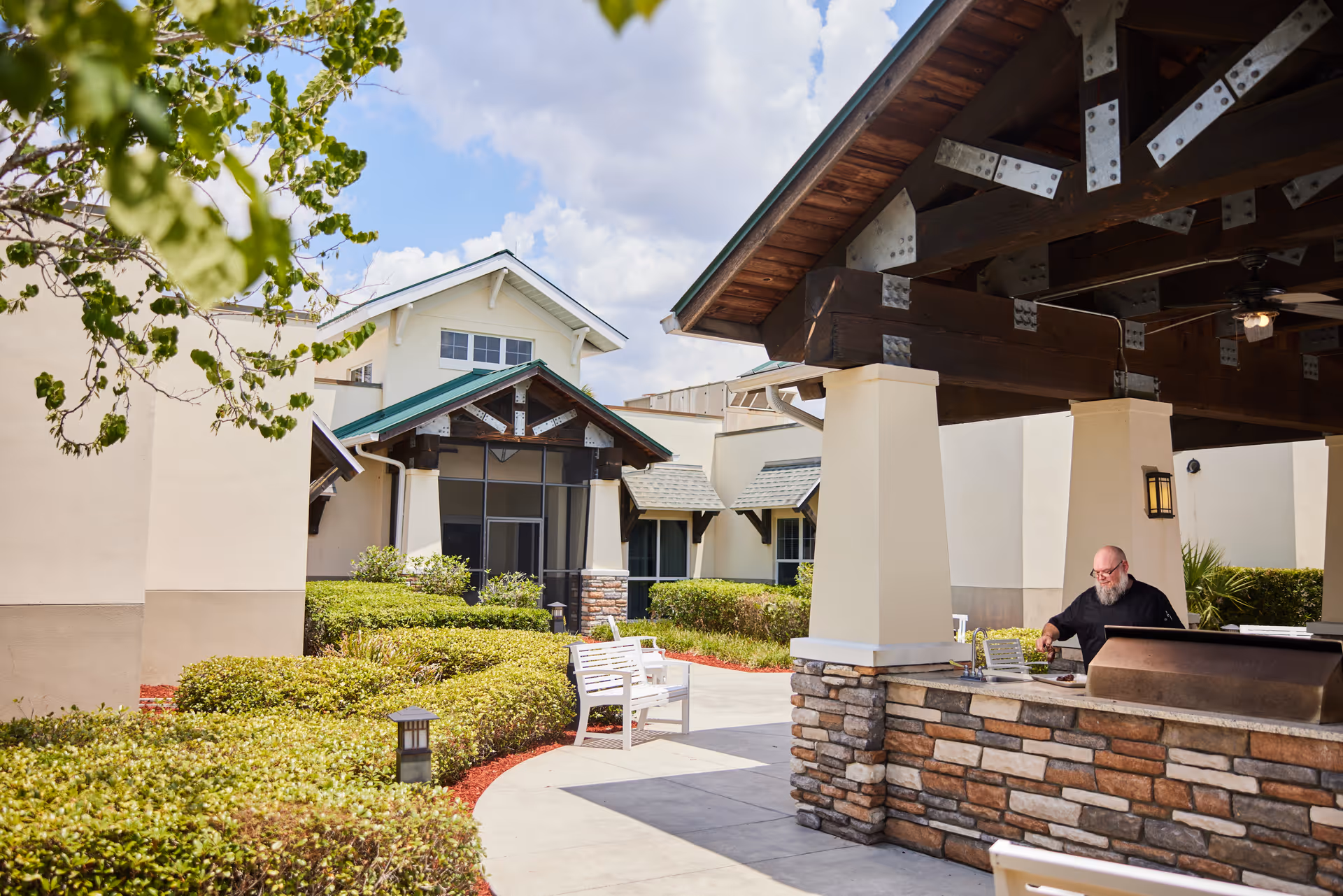 Outdoor patio area at Villages Rehab & Nursing Center with a man grilling food under a covered pavilion. The area features stone and wood construction, surrounded by bushes and greenery, with a white bench along the pathway leading to the building.