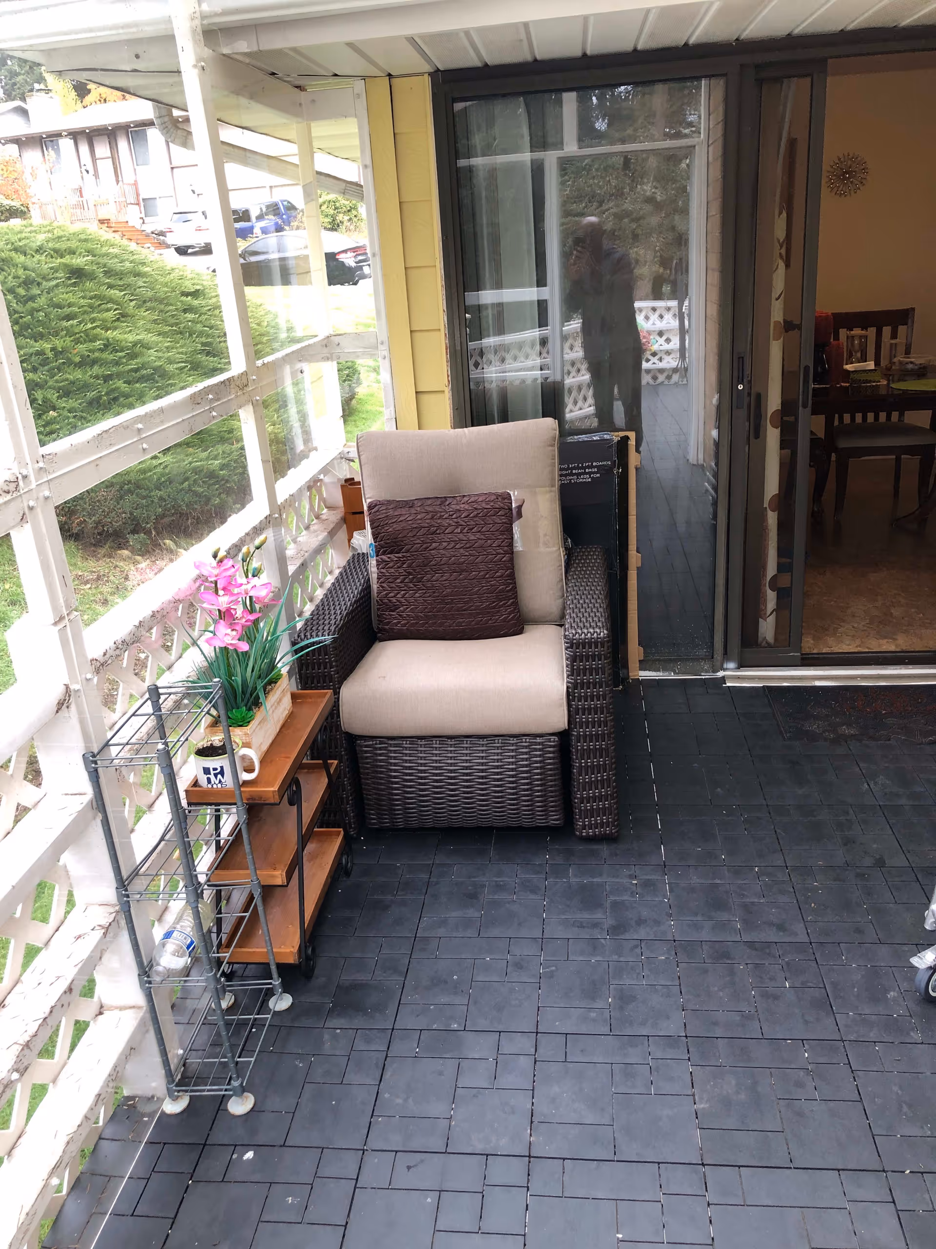 A small outdoor patio area with a cushioned wicker armchair featuring a brown pillow. Next to the chair is a small wooden shelf with pink flowers and a white mug on it. The patio has black tiled flooring and is enclosed with white lattice fencing. A sliding glass door leads inside to a dining area with a table and chairs. Green bushes and parked cars are visible outside the patio.