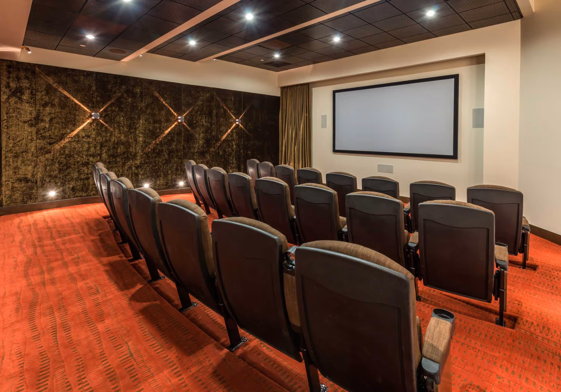 A small theater room with three rows of cushioned seats facing a large blank projection screen. The room has a red carpet with a patterned design, dark brown padded walls on one side with small lights, and a ceiling with recessed lighting.