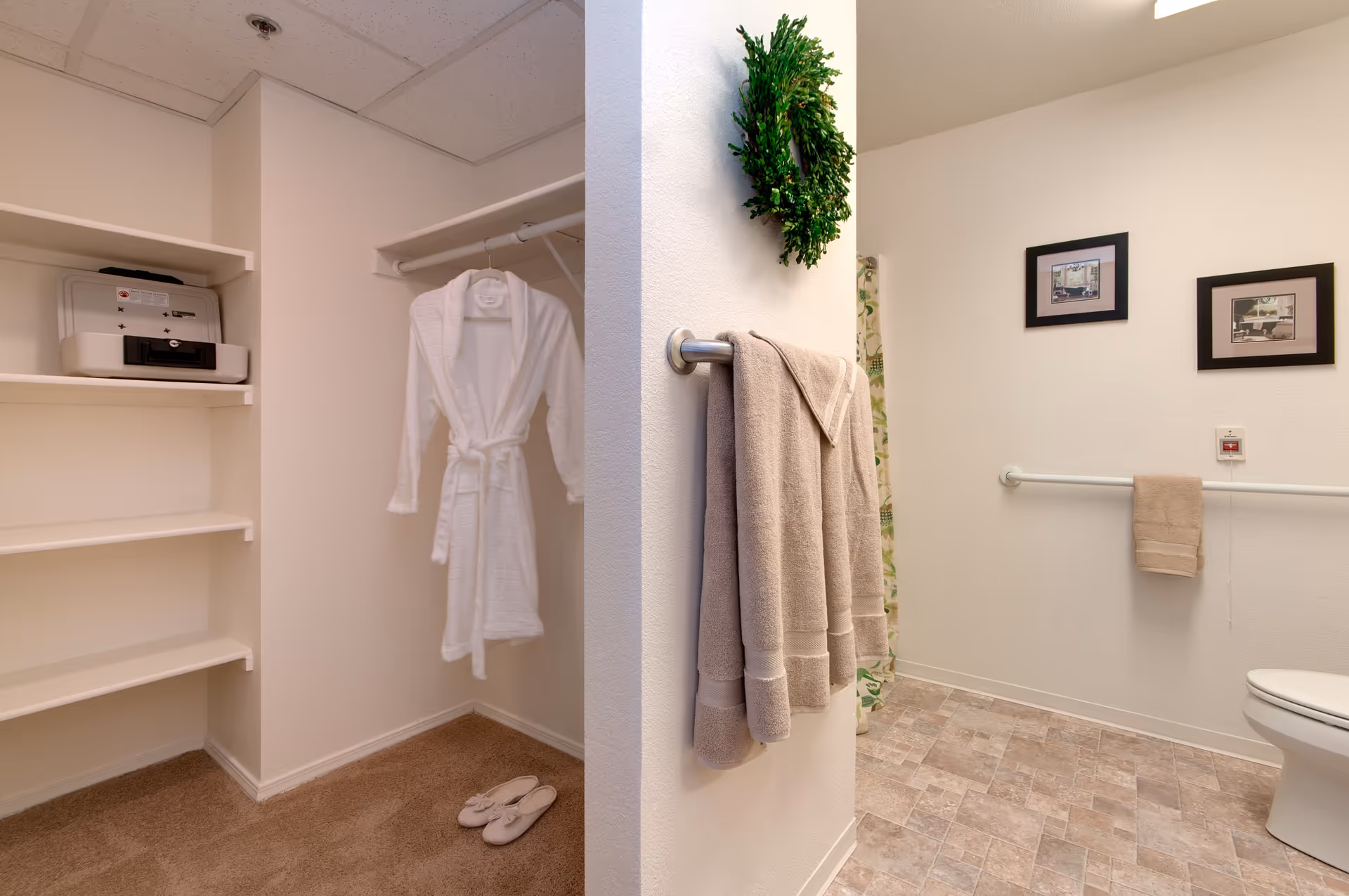 A bathroom and closet area in a senior living facility. The closet has white shelves, a hanging white bathrobe, and slippers on the carpeted floor. The bathroom features beige tiled flooring, a toilet, a towel rack with beige towels, a green wreath hanging on the wall, and two framed pictures. There is also a grab bar on the wall near the toilet.