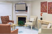 A cozy living room area with a fireplace, a mounted flat-screen TV above the fireplace, two beige armchairs, a patterned armchair, a small round wooden table with magazines, a wooden console table with a vase of flowers, and two abstract paintings on the wall.