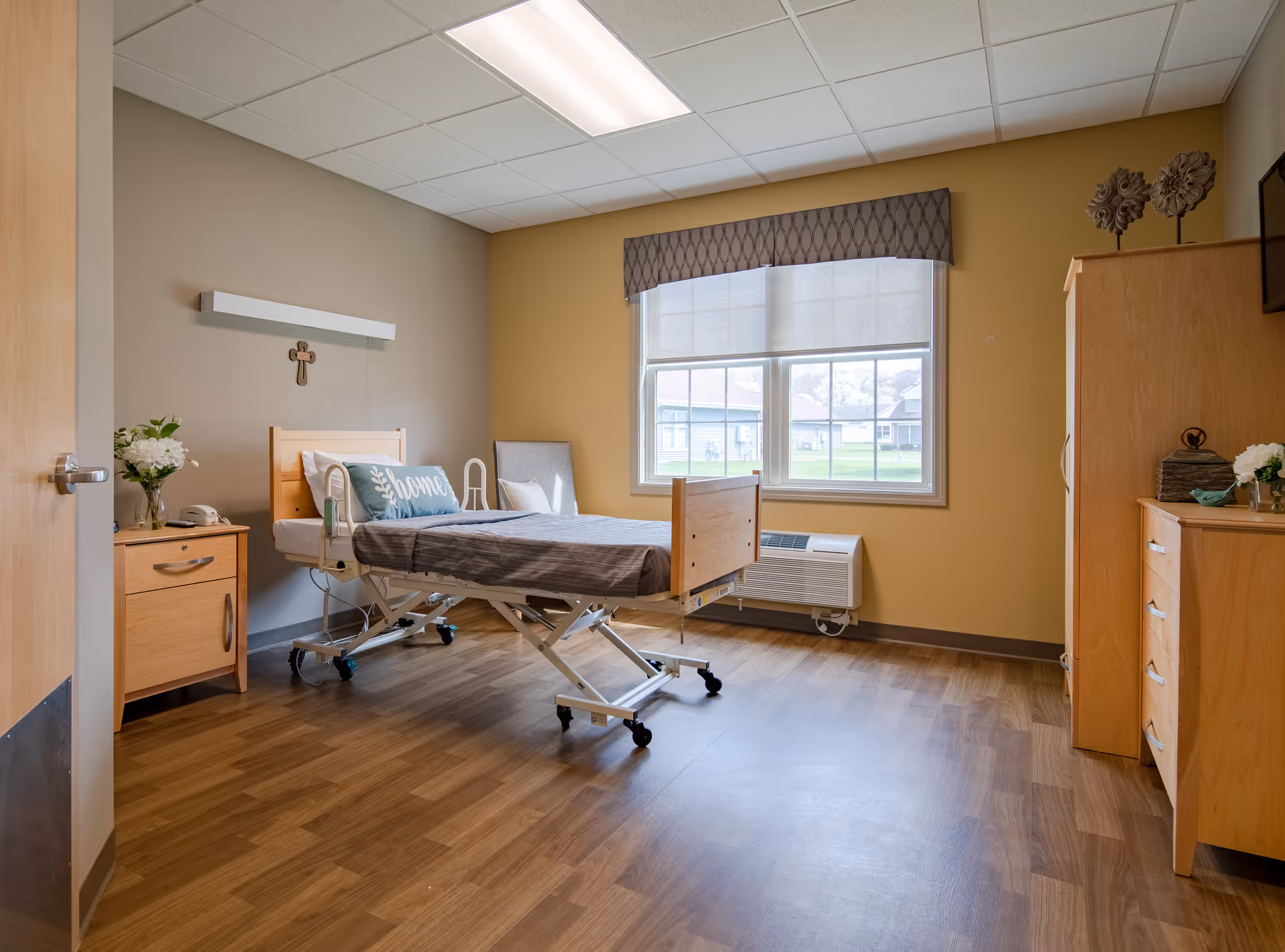 A clean and well-lit bedroom in a senior living facility with a hospital-style adjustable bed, a pillow with the word 'home' on it, wooden furniture including a nightstand and a dresser, a window with a valance, and decorative items such as flowers and wall art.