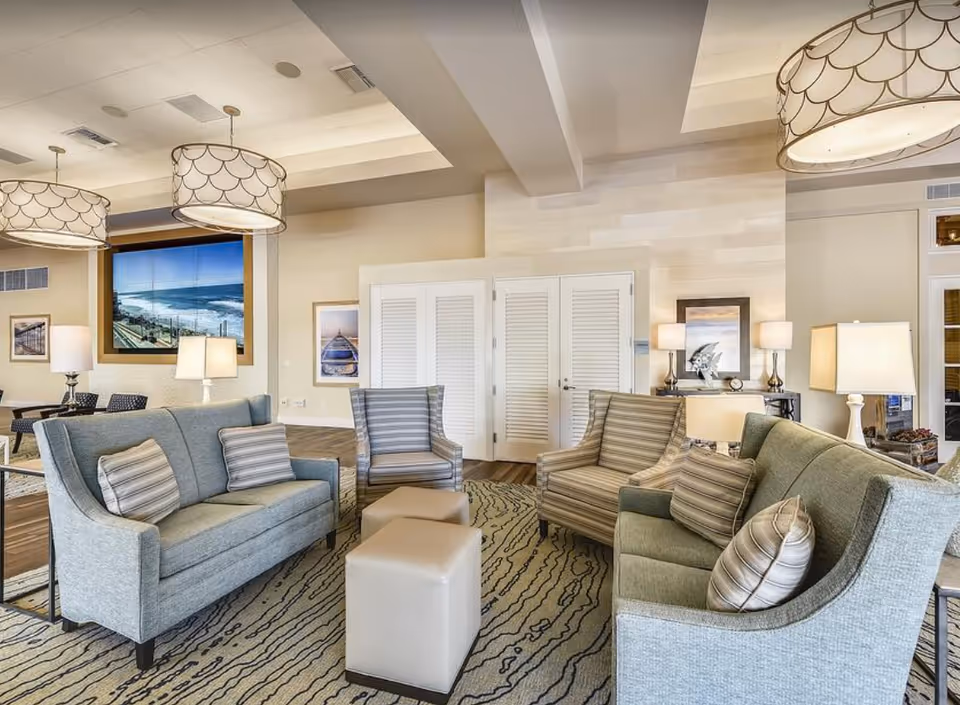 Bright coastal-themed communal lounge with sofas, striped armchairs, ottomans, table lamps, and a large wall-mounted ocean scene.