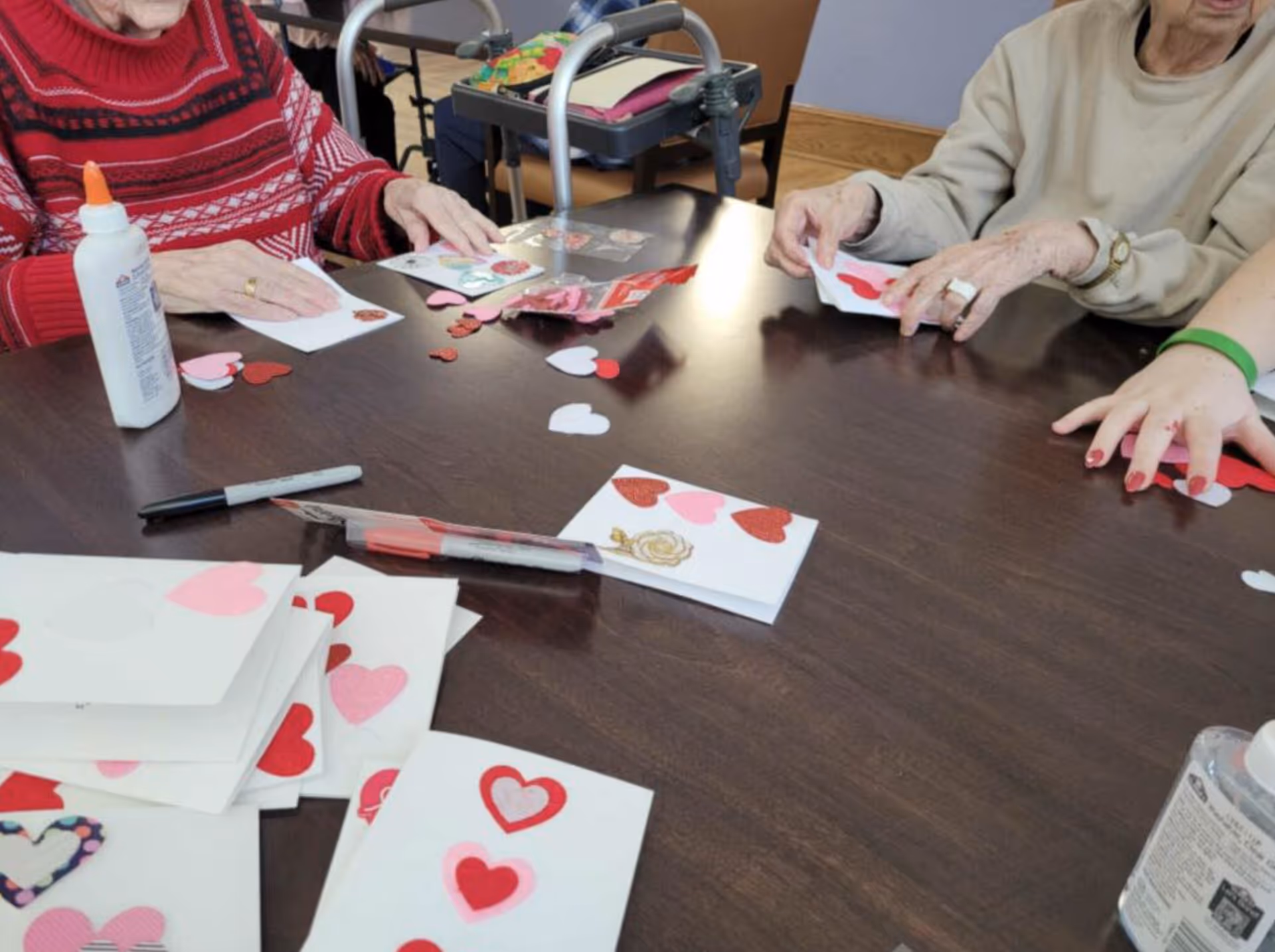 Several elderly individuals sitting around a dark wooden table engaged in a craft activity, decorating white cards with red and pink heart-shaped cutouts. Various craft supplies including glue, markers, and additional heart cutouts are scattered on the table.