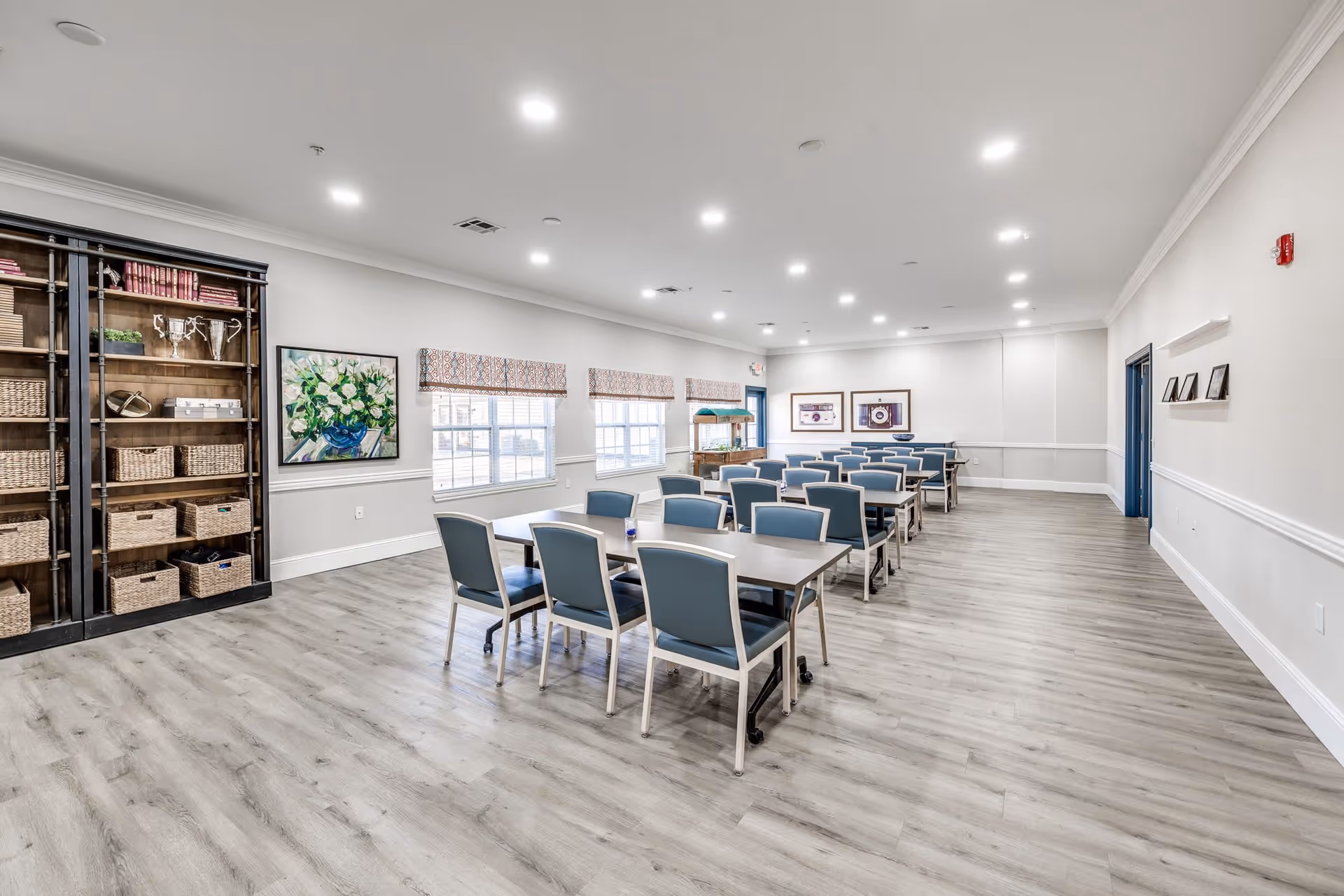 A spacious, well-lit room with multiple tables and blue chairs arranged in rows, large windows with patterned valances, a tall bookshelf filled with baskets and decorative items, and framed artwork on the walls.