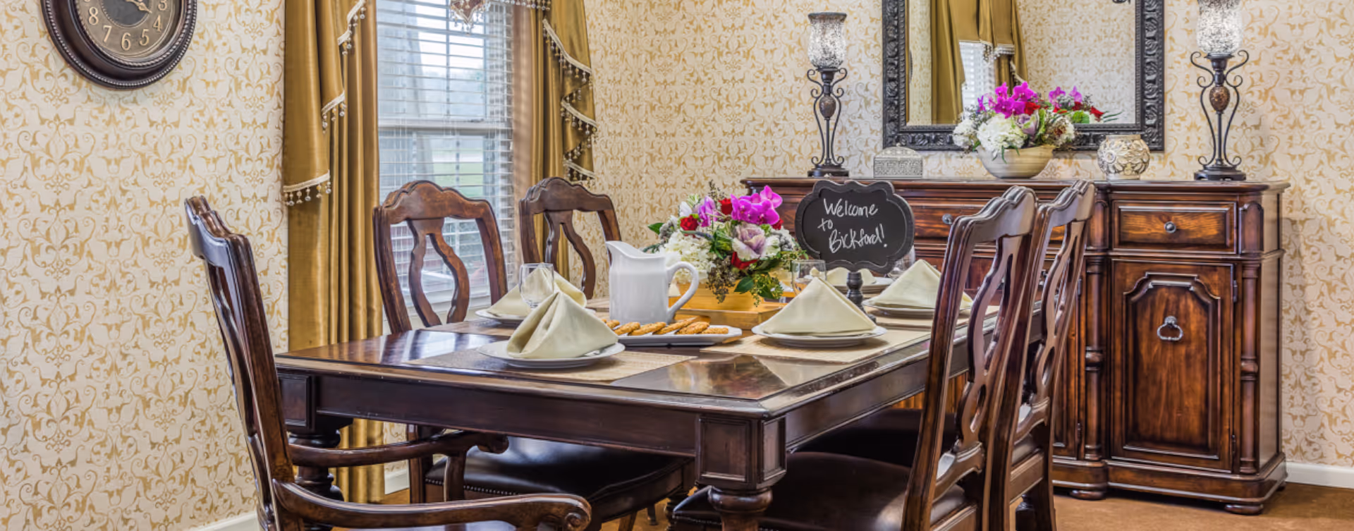 A formal dining room with a dark wooden dining table set for four with folded napkins, a white pitcher, and a plate of cookies. The room has patterned wallpaper, gold curtains, a wall clock, and a wooden sideboard with decorative lamps, a mirror, and flower arrangements including a small chalkboard sign that says 'Welcome to Bickford!'