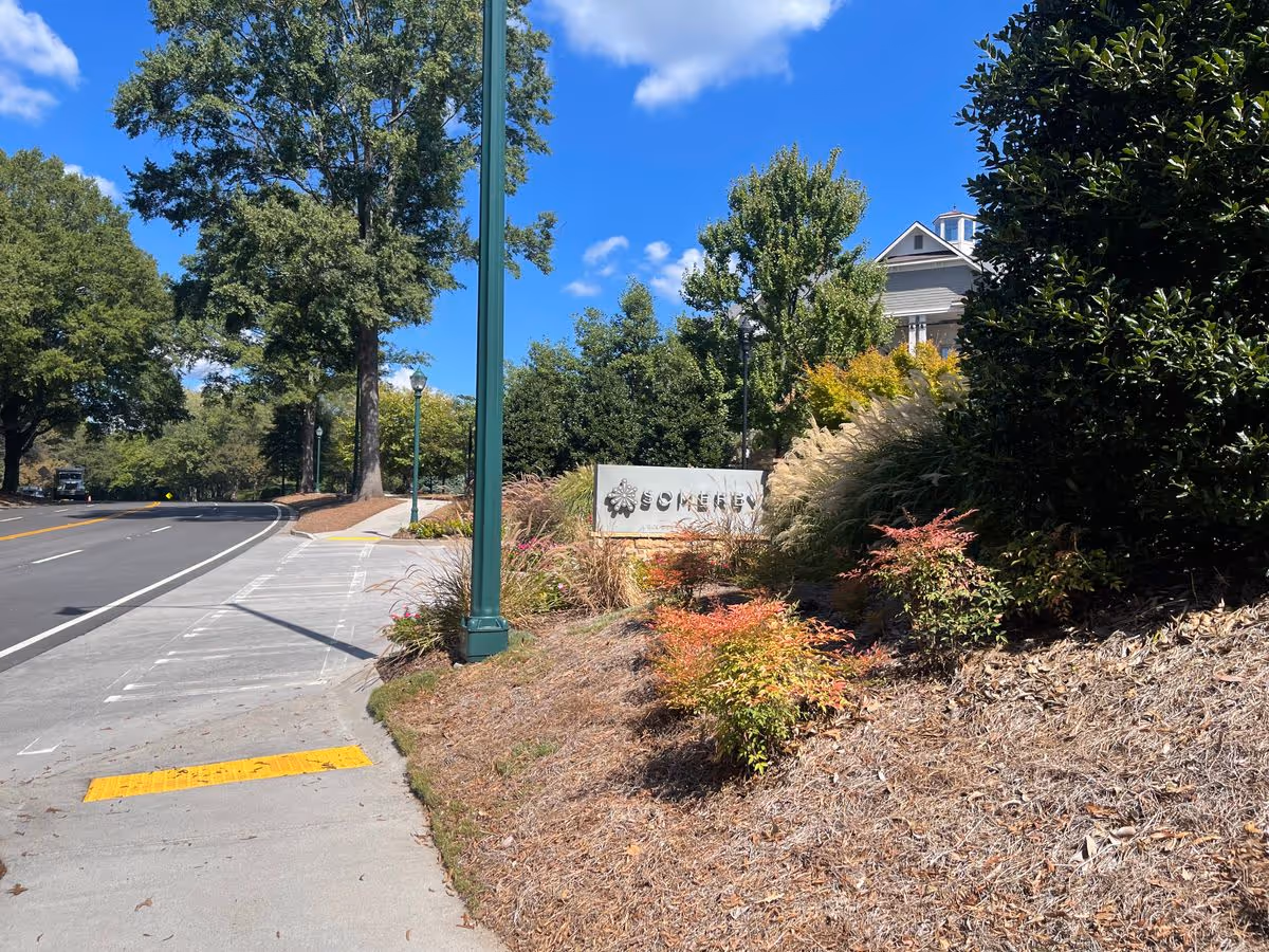 Sidewalk and roadway passing a landscaped entrance sign for Somerby senior living with trees, lamp posts and a building in the background under a blue sky.