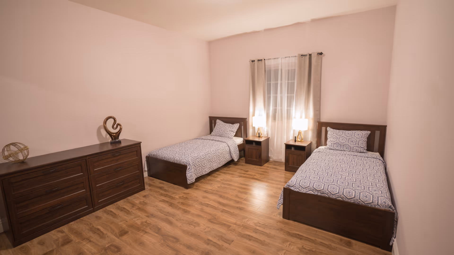 A bedroom with two single beds featuring matching patterned bedding. Between the beds is a window with sheer and beige curtains, flanked by two nightstands each with a lamp. On the left side of the room is a wooden dresser with decorative items on top. The floor is wooden and the walls are painted light pink.