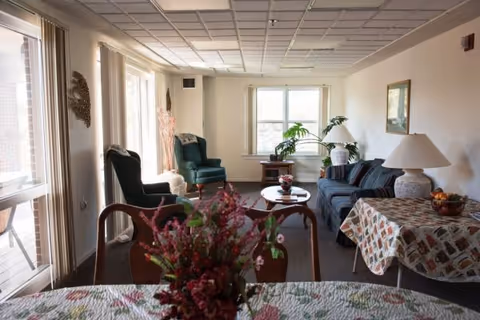 Sunlit common living area with sofas, armchairs, side tables and a dining table with a floral centerpiece.