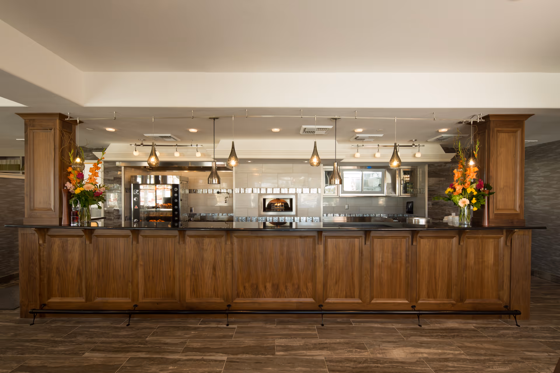 A modern kitchen area with a long wooden counter in the foreground, pendant lights hanging from the ceiling, and a tiled backsplash with a built-in oven and stainless steel appliances. There are two vases with colorful flowers on each end of the counter.