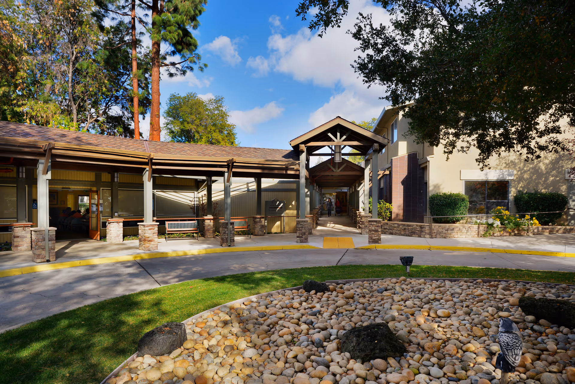 Exterior view of a senior living facility with a covered walkway supported by stone pillars, surrounded by trees and landscaping including a circular rock garden in the foreground under a blue sky with some clouds.