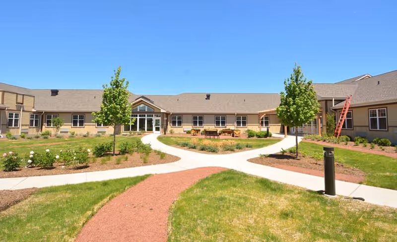 Exterior view of Silverado St. Charles Memory Care Community showing a single-story building with beige siding and multiple windows surrounding a landscaped courtyard with green grass, small trees, flower beds, and paved walkways under a clear blue sky.