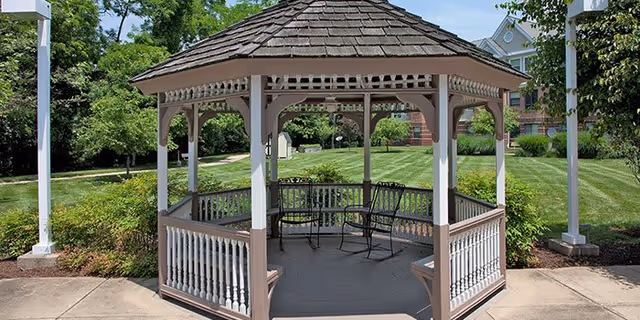 A wooden gazebo with a shingled roof and white railings situated on a paved area surrounded by green grass, trees, and shrubs. Two metal chairs and a small table are placed inside the gazebo. In the background, there is a large brick building partially visible.