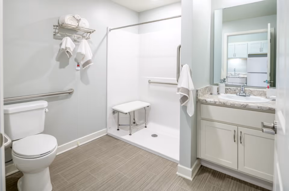 A clean and accessible bathroom featuring a toilet, a walk-in shower with a foldable bench and grab bars, a towel rack with rolled towels, and a vanity with a sink and mirror. The bathroom has light-colored walls and tiled flooring.