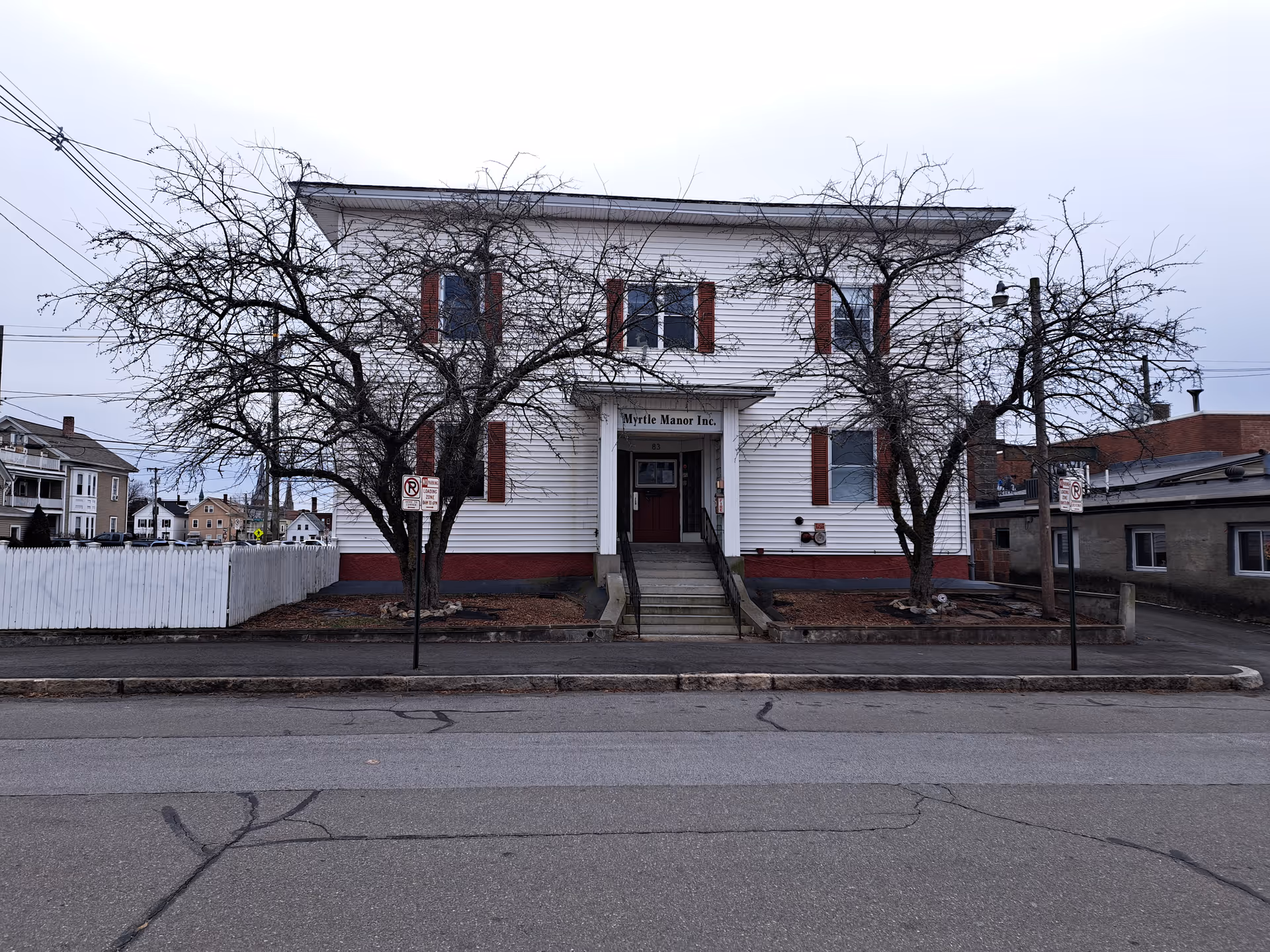 Front exterior view of a two-story white building with red shutters and a small staircase leading to the entrance. The building has a sign above the door that reads 'Myrtle Manor Inc'. Two leafless trees are in front of the building, and there are no people visible. The street and sidewalk are in the foreground.