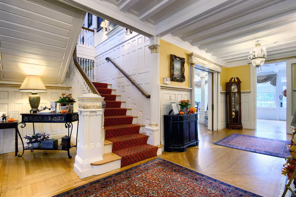 A bright and elegant interior hallway featuring a wooden staircase with a red carpet runner, white paneled walls, and decorative columns. The space includes ornate rugs on the wooden floor, a grandfather clock, a black side cabinet with flowers and small pumpkins, and a table with a lamp and additional decorations under the staircase. The ceiling has exposed beams and a chandelier, with doorways leading to other rooms.