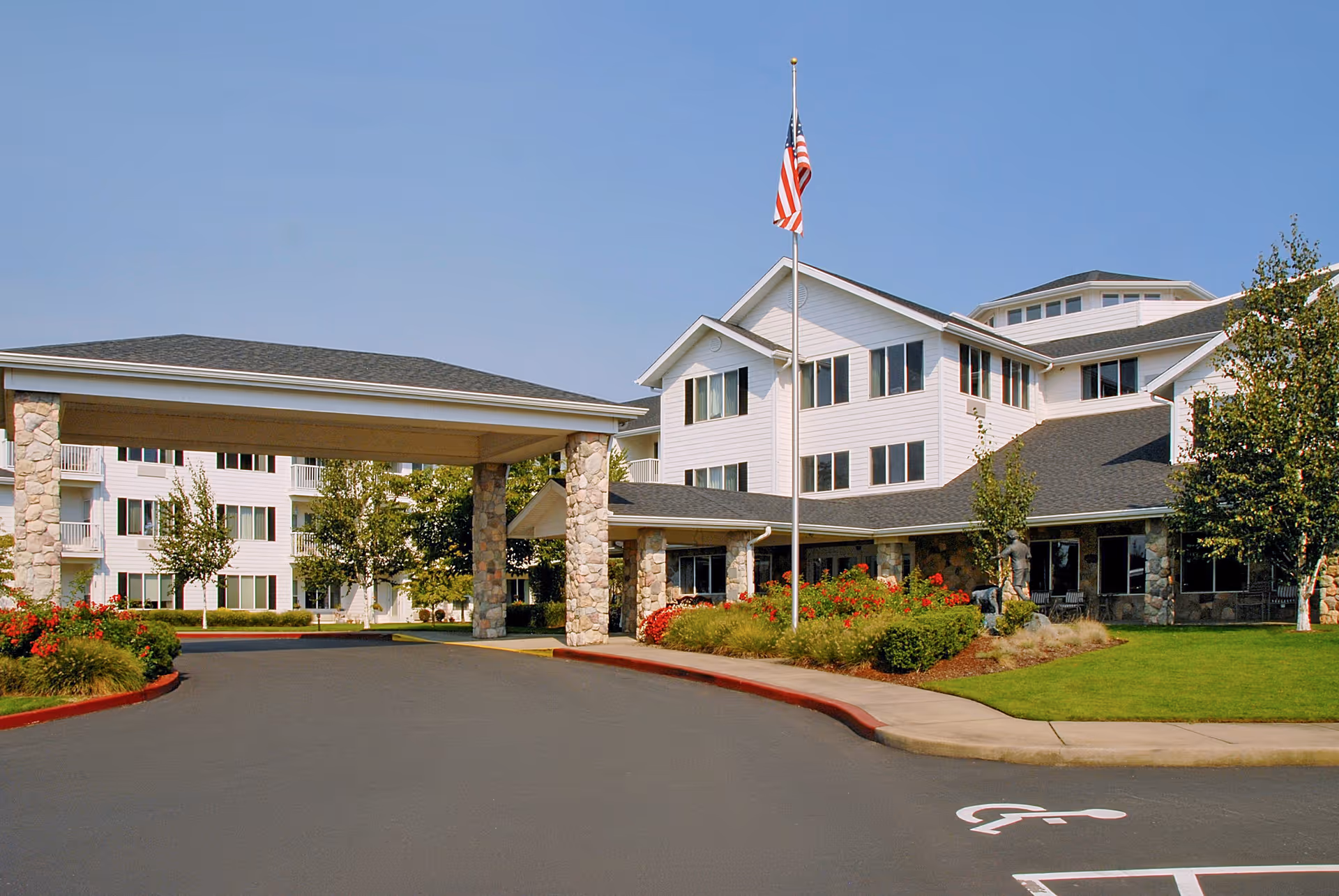 Exterior view of a senior living facility named Solista Newberg featuring a large covered entrance supported by stone pillars, a well-maintained driveway with a handicap parking space, landscaped greenery with bushes and trees, and an American flag on a flagpole in front of the building under a clear blue sky.