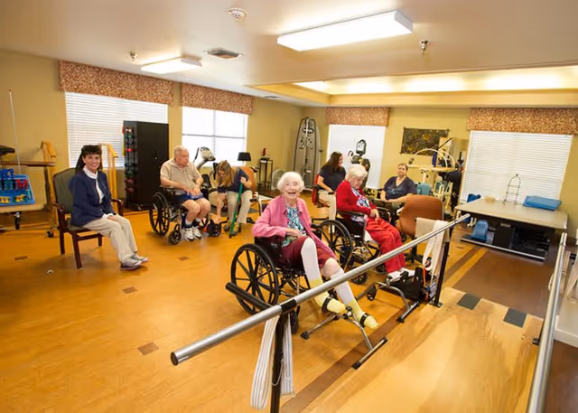 A group of elderly individuals, some in wheelchairs, participating in a physical therapy or rehabilitation session in a well-lit room with exercise equipment and parallel bars. A few caregivers or staff members are assisting them.