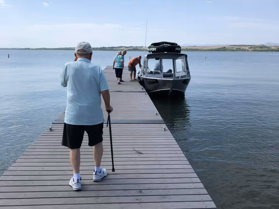 An older person using a cane walks along a wooden dock toward a small boat while two other people stand near the boat on a calm lake.