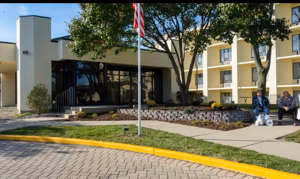 Exterior view of High Point Residence Kankakee Supportive Living & Memory Care facility showing the entrance area with a flagpole, landscaped garden beds, and two elderly people sitting on a bench near the building. The building has multiple windows and a light yellow facade.
